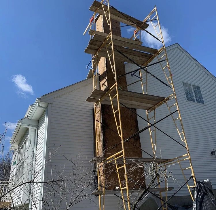 Chimney repair: Scaffolding surrounds a damaged chimney on a two-story white house against a blue sky.