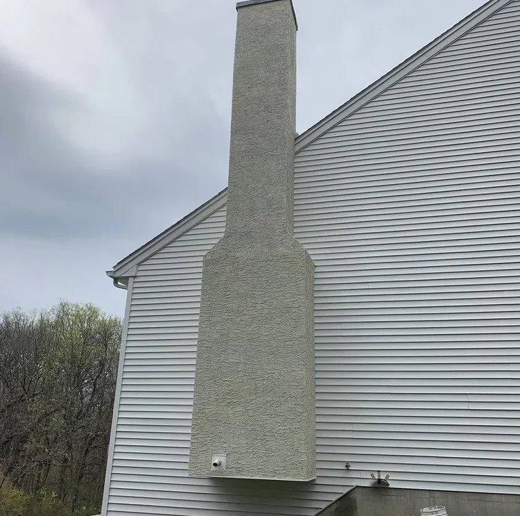 Tall, light gray stucco chimney on the side of a white-sided house with a gray roof; overcast sky.
