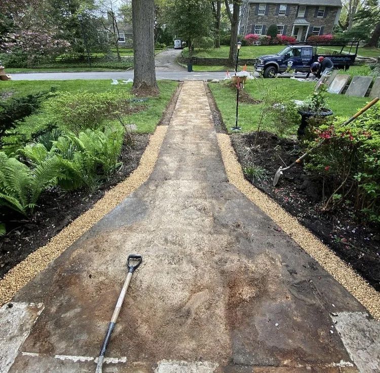 A pathway being renovated with gravel edging. Shovel lies on the weathered surface. A truck and workers are in the background.