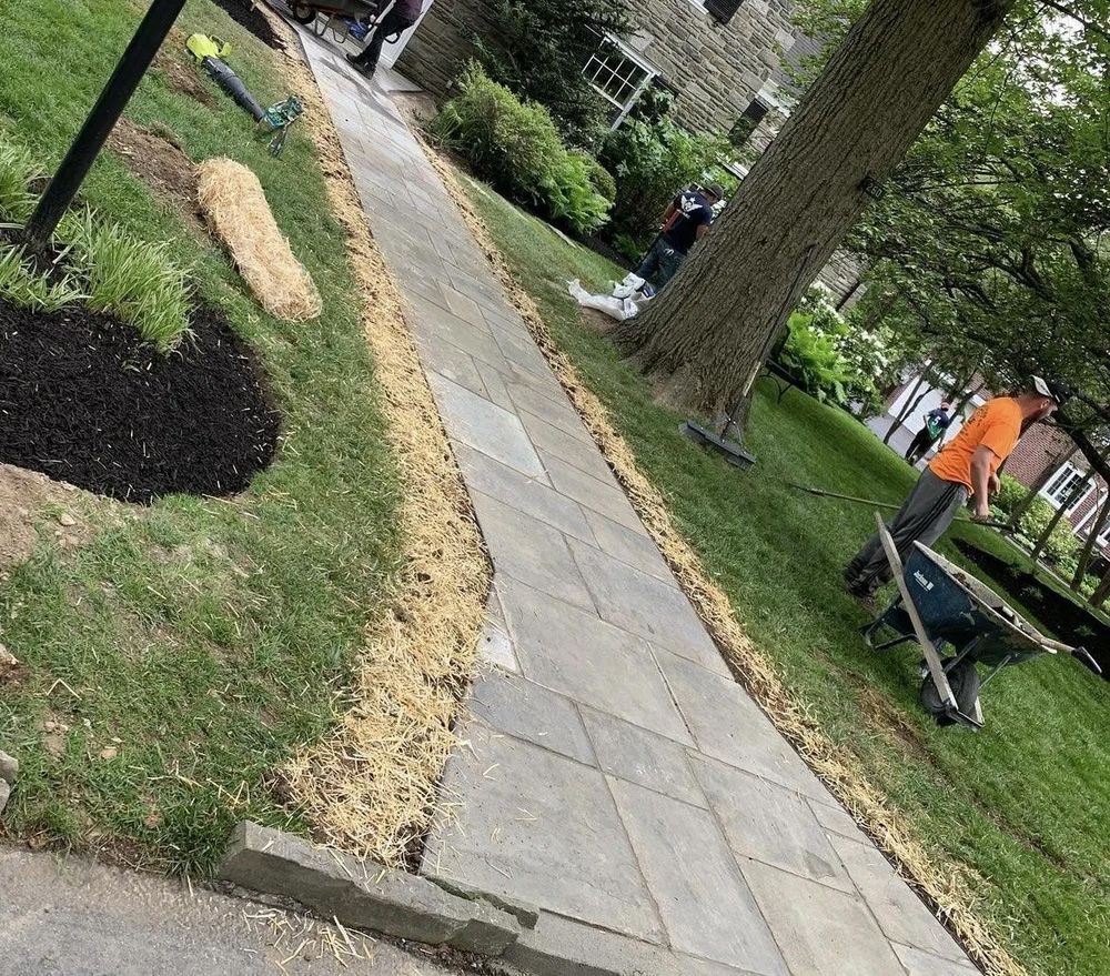 Workers edging a stone walkway, spreading straw along the side of the path next to grass and a tree.