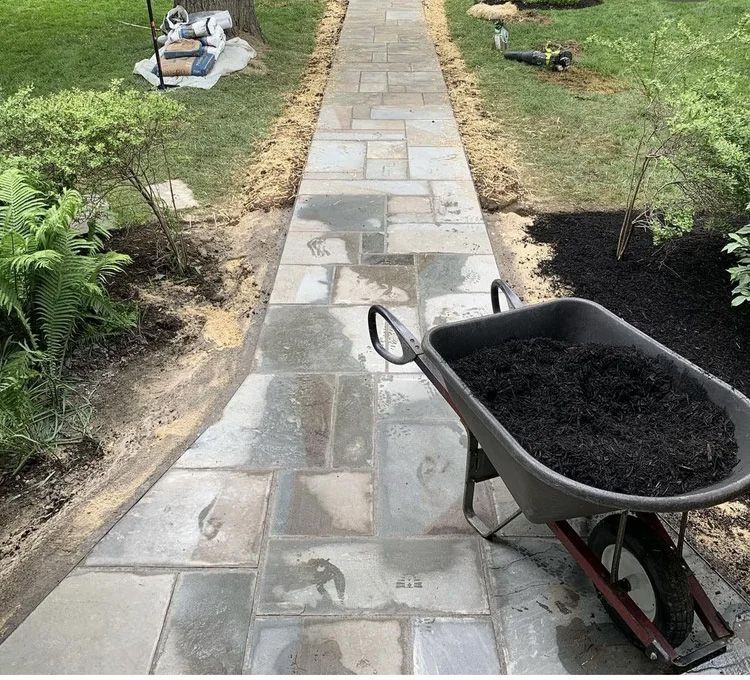 Stone walkway bordered by dirt, with a wheelbarrow full of mulch, surrounded by green grass and shrubs.