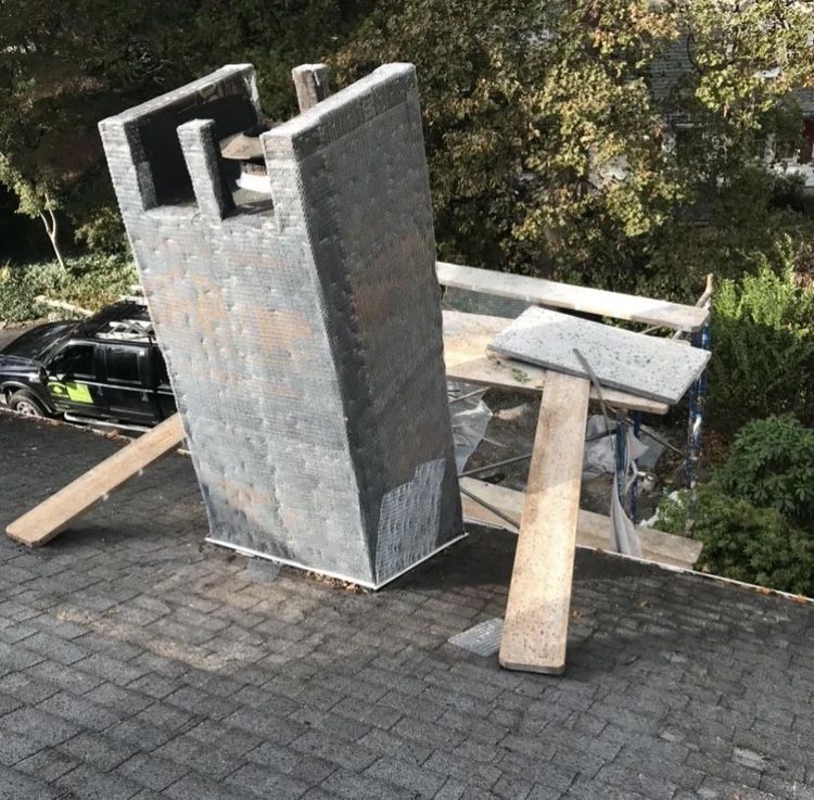 A large, gray chimney on a dark roof, propped up with wooden planks, near a black truck.