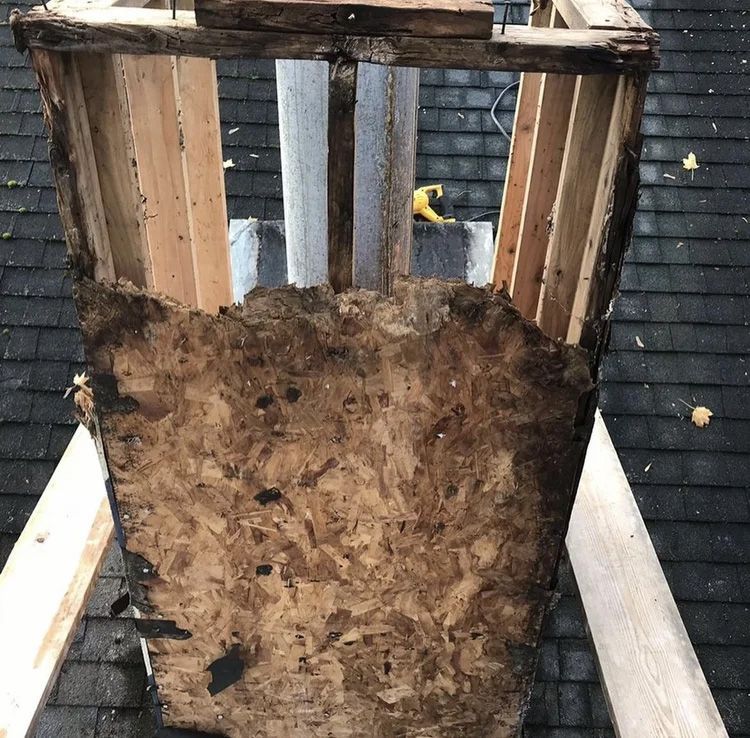 Partially dismantled chimney on a shingled roof, showing wood framing and weathered particle board.