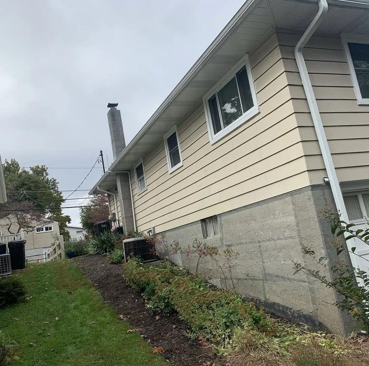 Beige house on a sloped lot with a concrete foundation, chimney, and gutters, overcast sky.