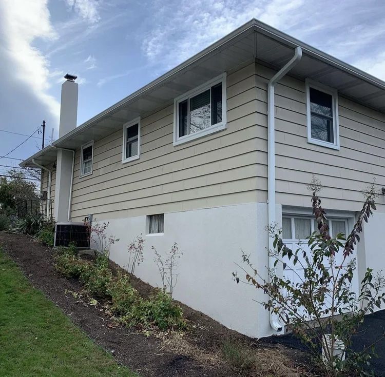 Two-story house with tan siding and white trim on a sloped lot under a cloudy sky.