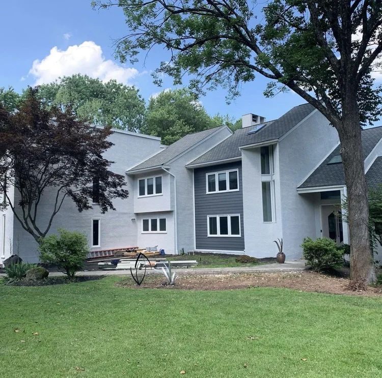 Two-story house with gray siding, white trim, and a dark gray accent wall. Lush green lawn and mature trees.