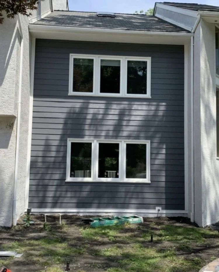 Dark gray siding with white-framed windows on a building exterior. Green grass and ground-level construction area.
