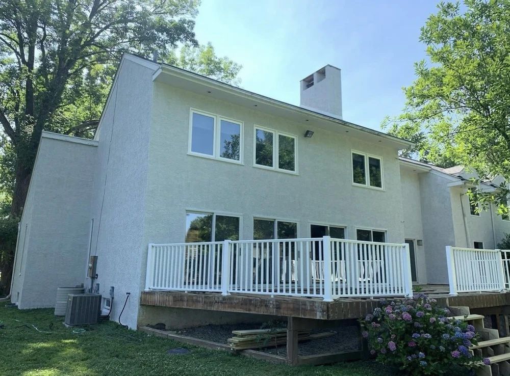 Two-story house with a white deck and light-colored stucco exterior; green trees and blue sky.