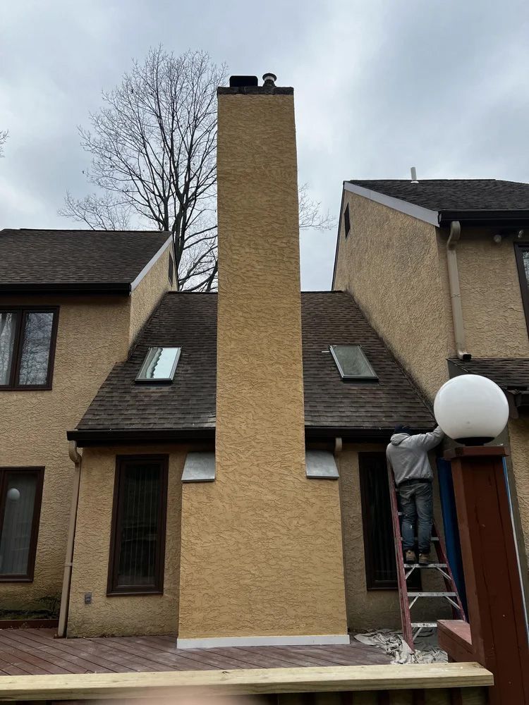 Tall yellow chimney on a home with a person on a ladder, overcast sky.