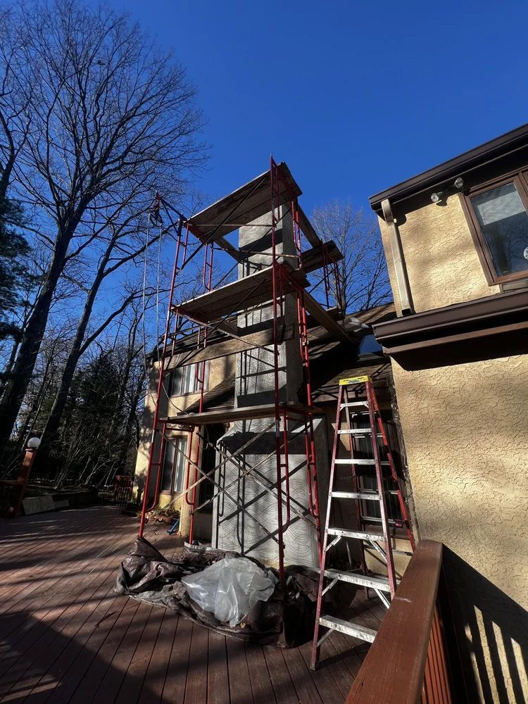 Scaffolding surrounds a chimney on a house exterior; ladder nearby.