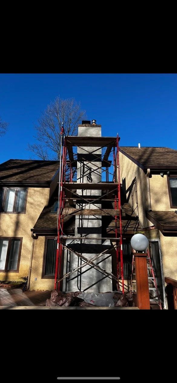 Chimney repair with scaffolding in front of a yellow house with a brown roof under a blue sky.