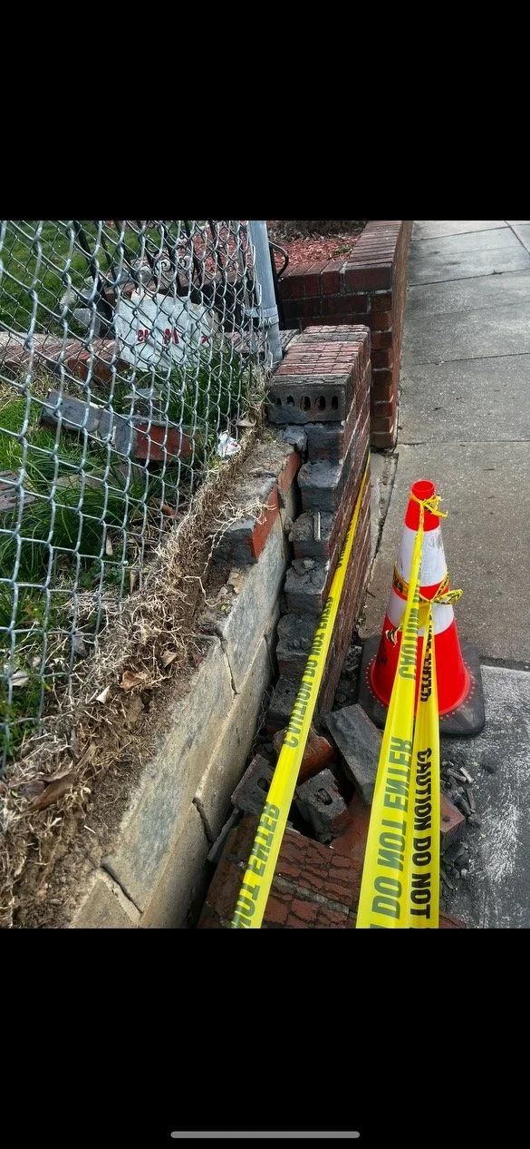 A brick wall with structural damage is cordoned off with caution tape and a traffic cone.