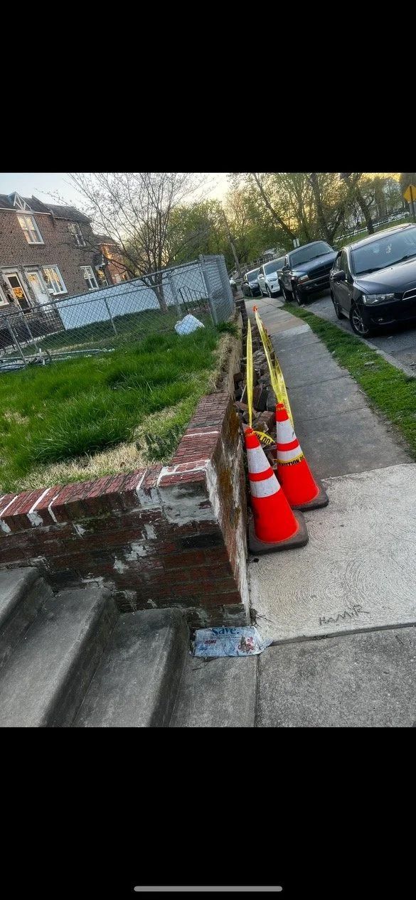 Two orange traffic cones on a sidewalk, next to a low brick wall and grass. Cars parked on the street.