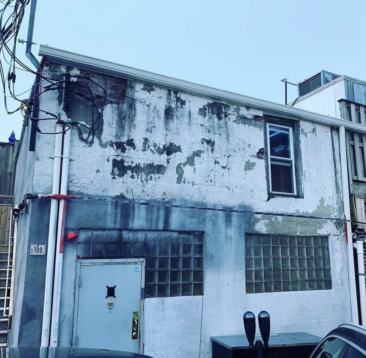 A dilapidated two-story building with peeling paint, a single window, and a glass-block entryway.