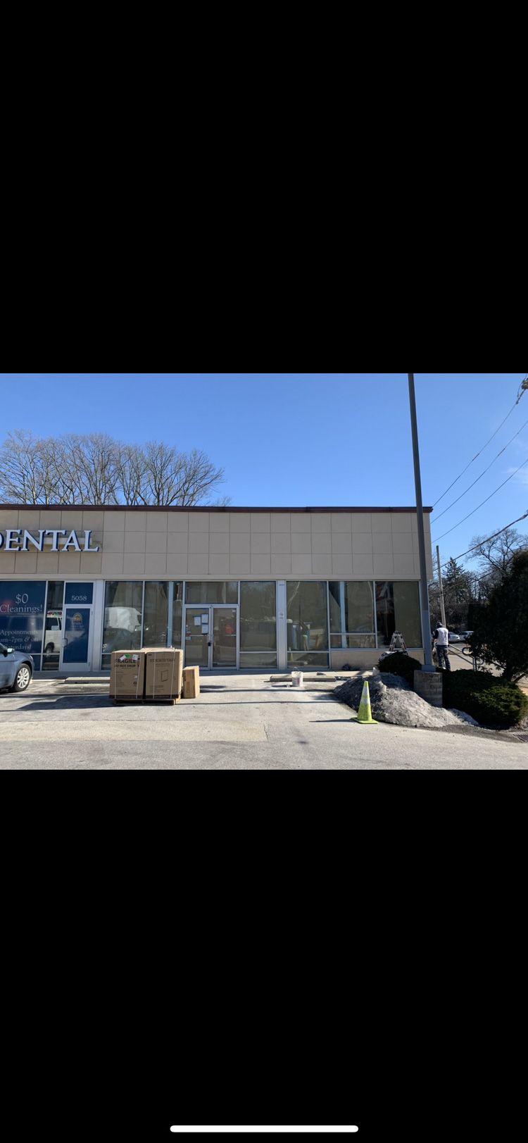Exterior of a storefront with large windows and tan facade. Boxes and construction materials in front. Bright blue sky.
