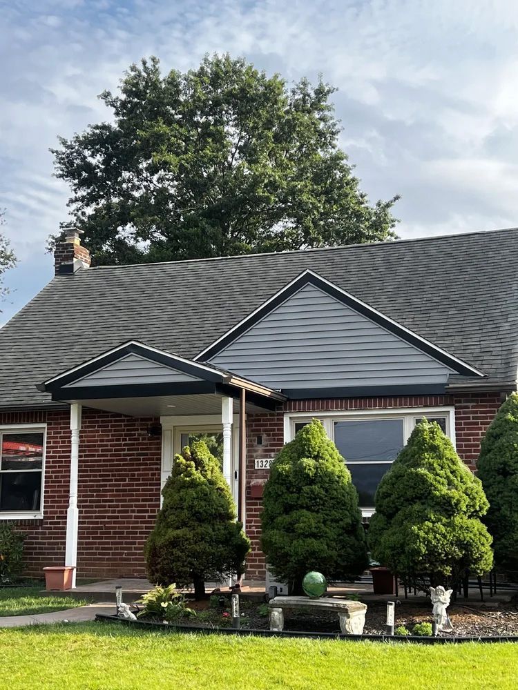 Brick house with dark roof and trim, green lawn, and trimmed evergreen shrubs. Cloudy sky.
