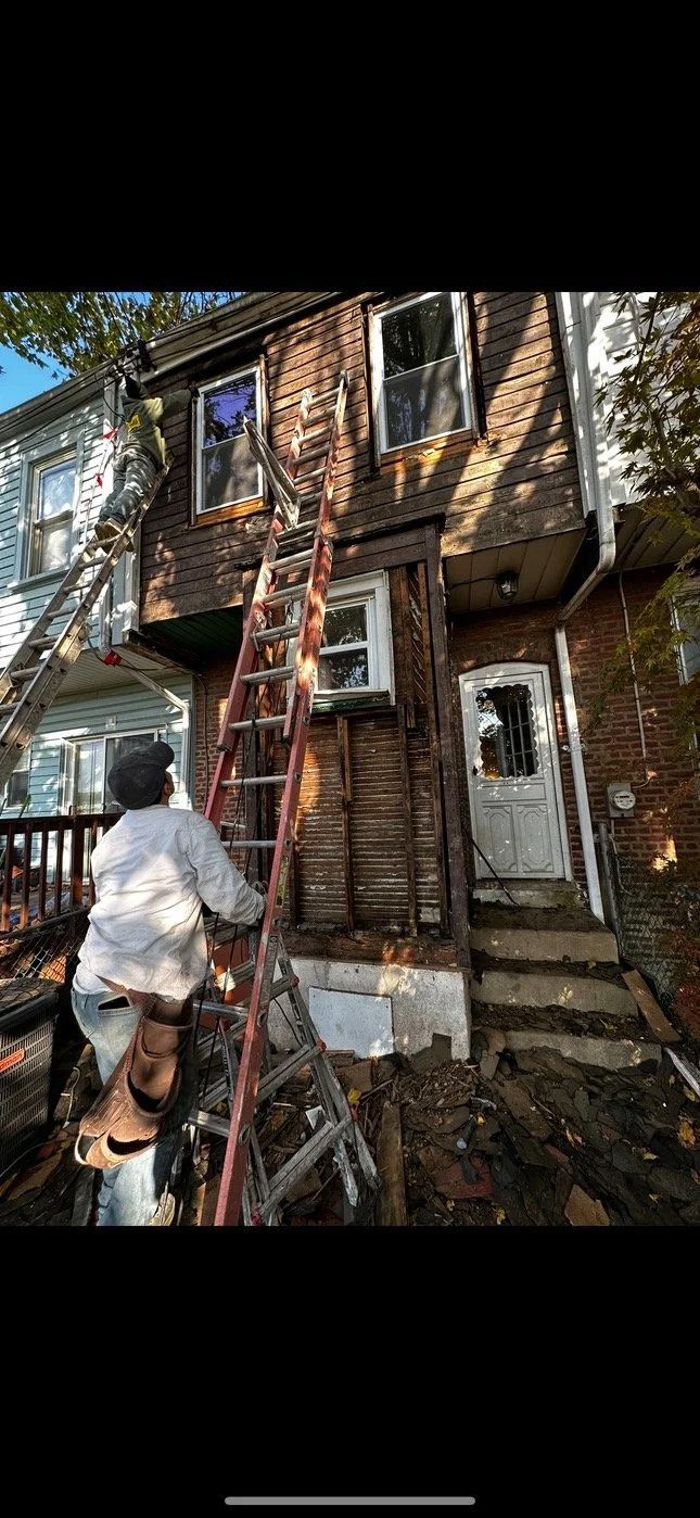 A person climbs a ladder to a weathered brick building with two windows.