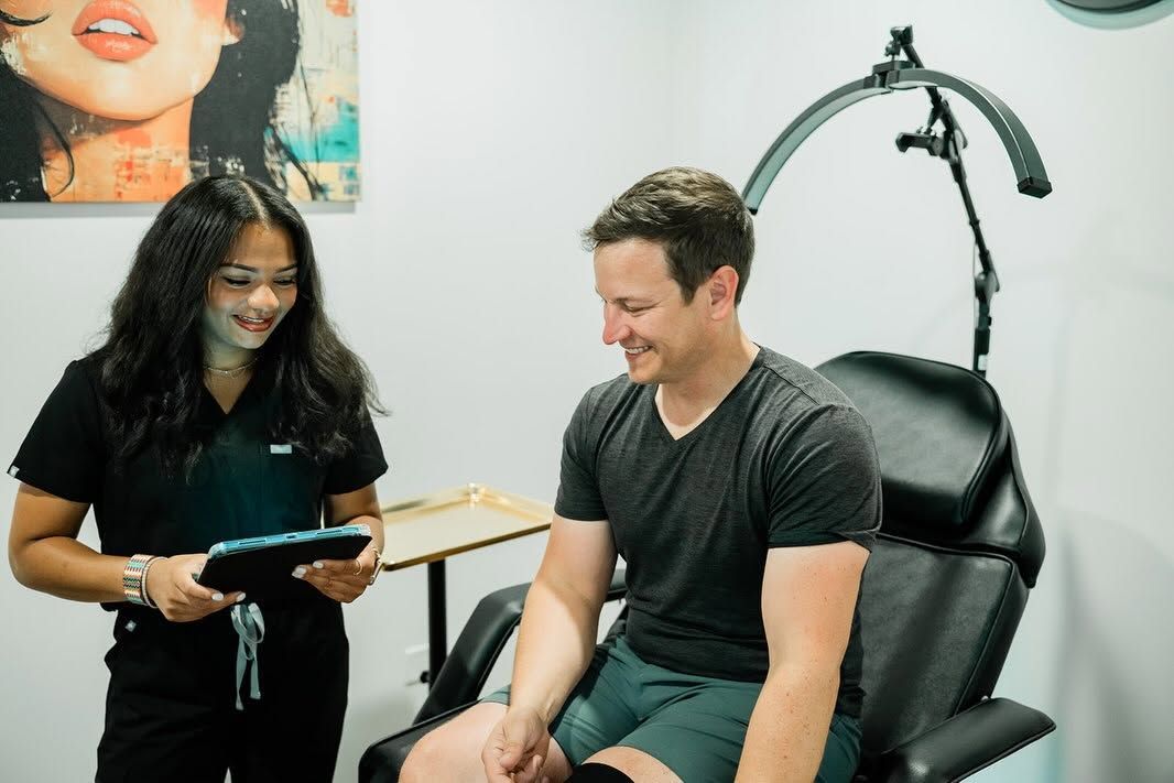 Two people smiling in a clinic, one standing with a tablet beside a reclining chair under a hooded lamp.