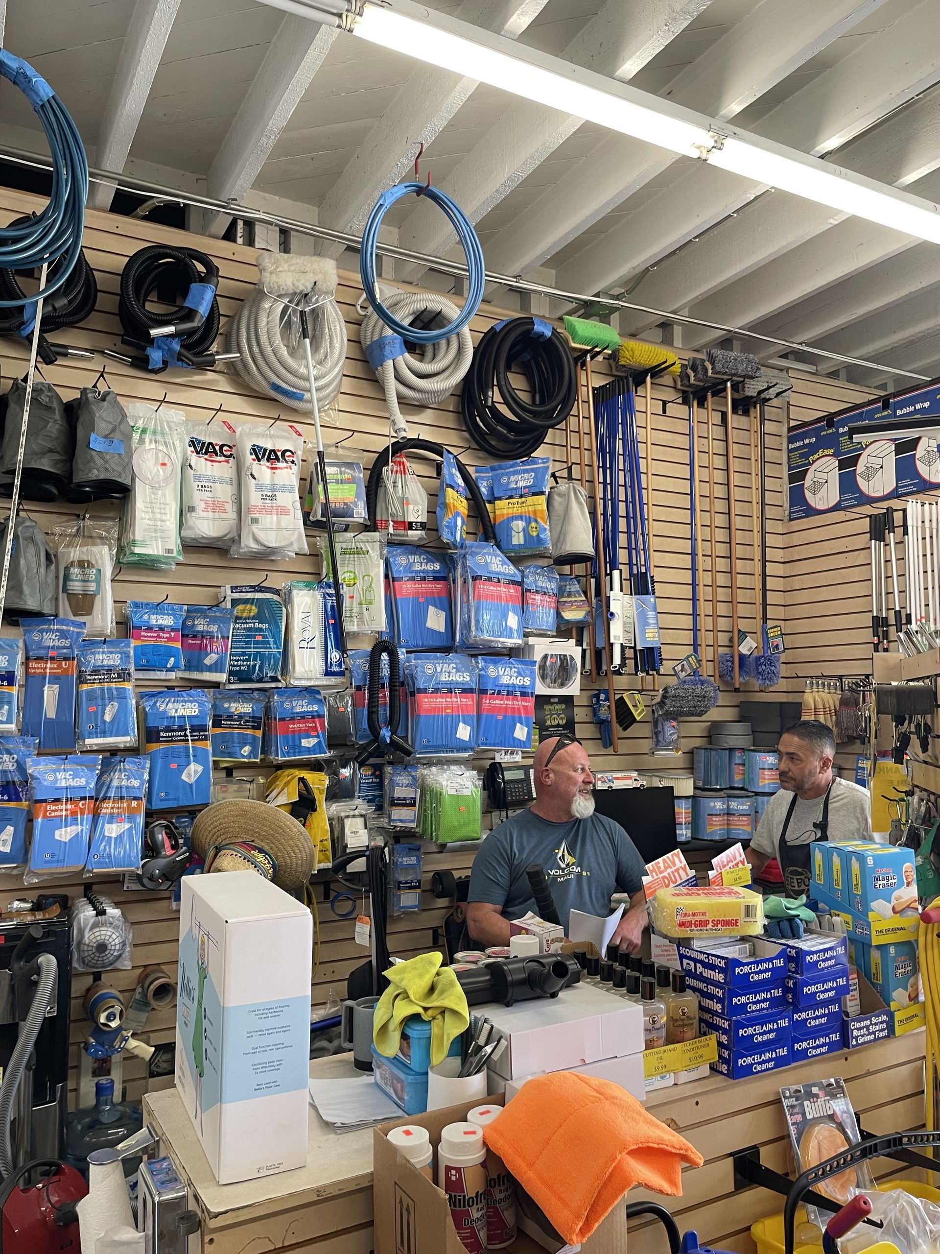 Store interior with two men at a checkout counter, surrounded by shelves of supplies; blue, white, and yellow dominate.