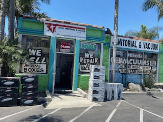 Local Janitorial & Vacuum store. Green and white building sells moving boxes and repairs vacuums. Sunny day.