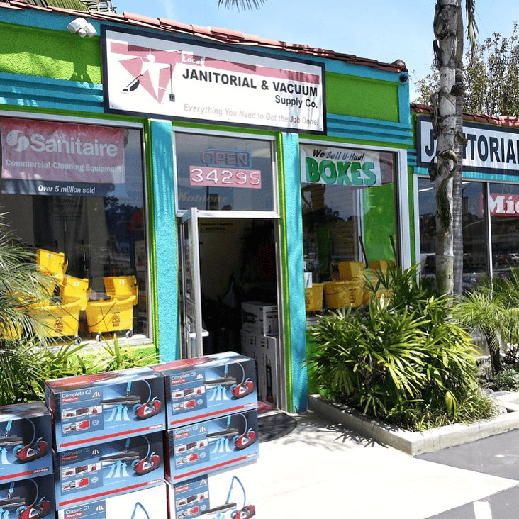 A green building: Local Janitorial & Vacuum store, sells boxes, and repairs vacuums. Boxes stacked outside.