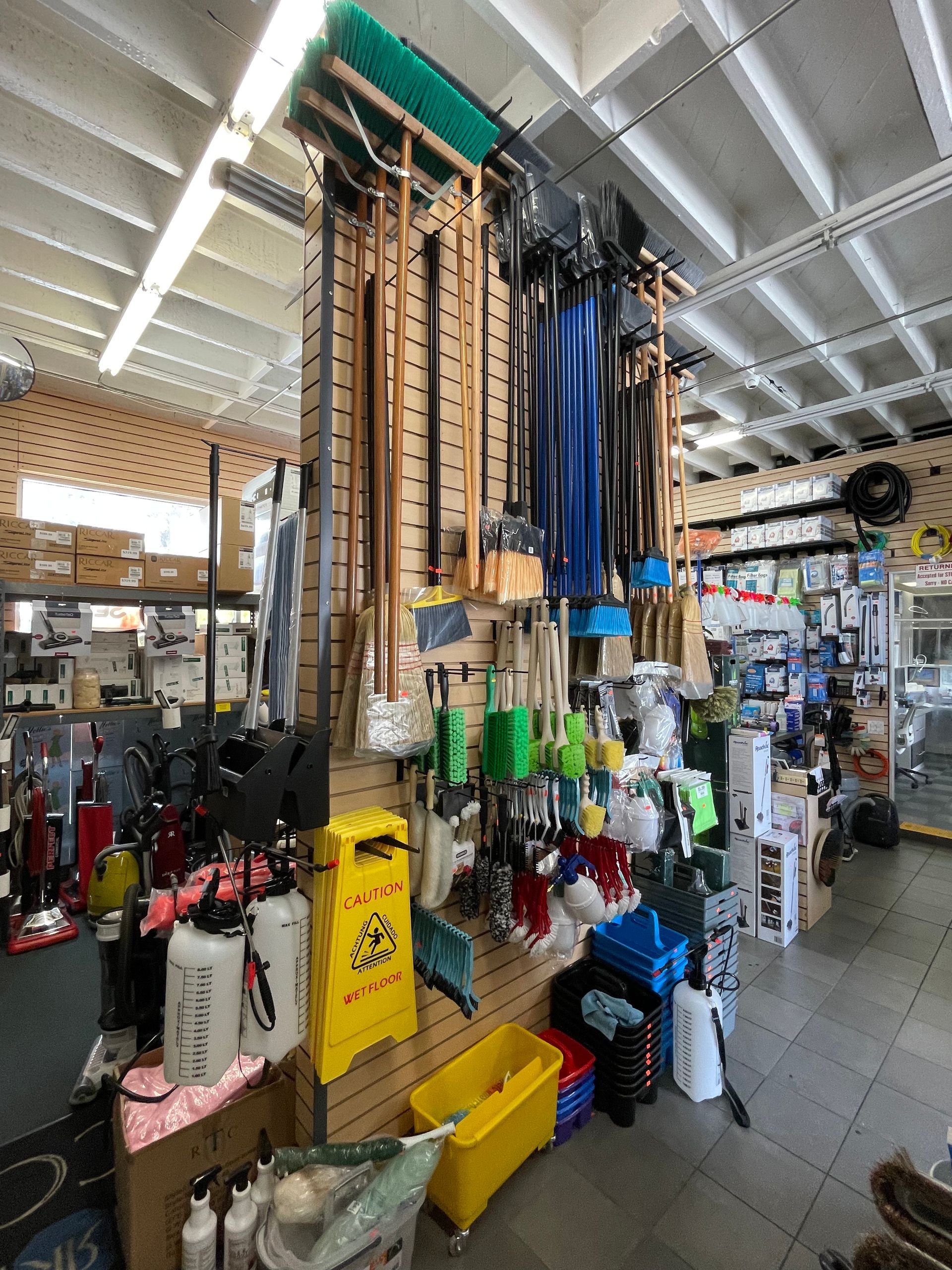 Cleaning supply store interior with brooms, mops, sprayers, and caution sign displayed on wall and shelves.