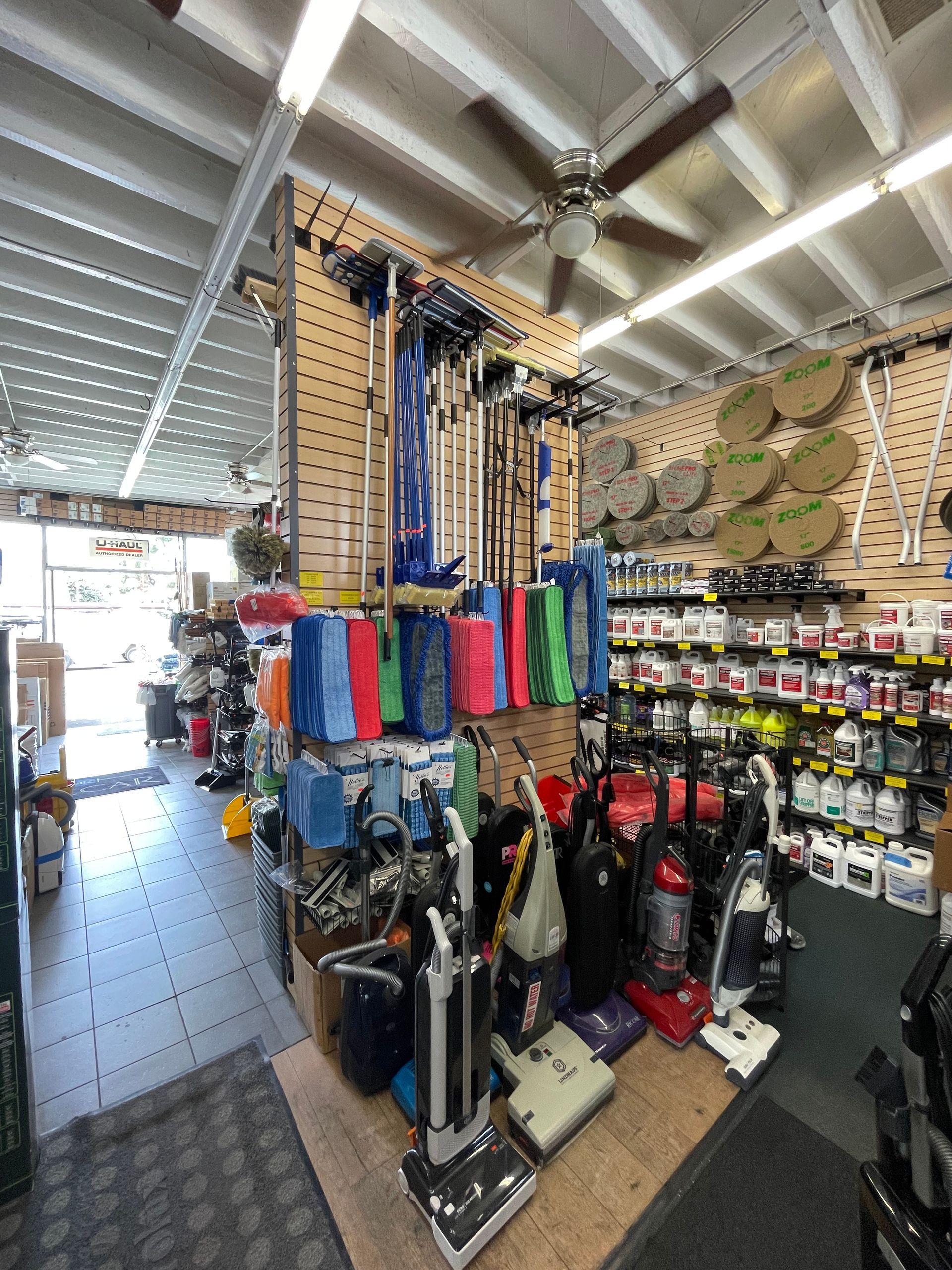 Inside a store, displaying cleaning supplies: vacuums, cloths, and chemicals. Wooden walls, ceiling fan.