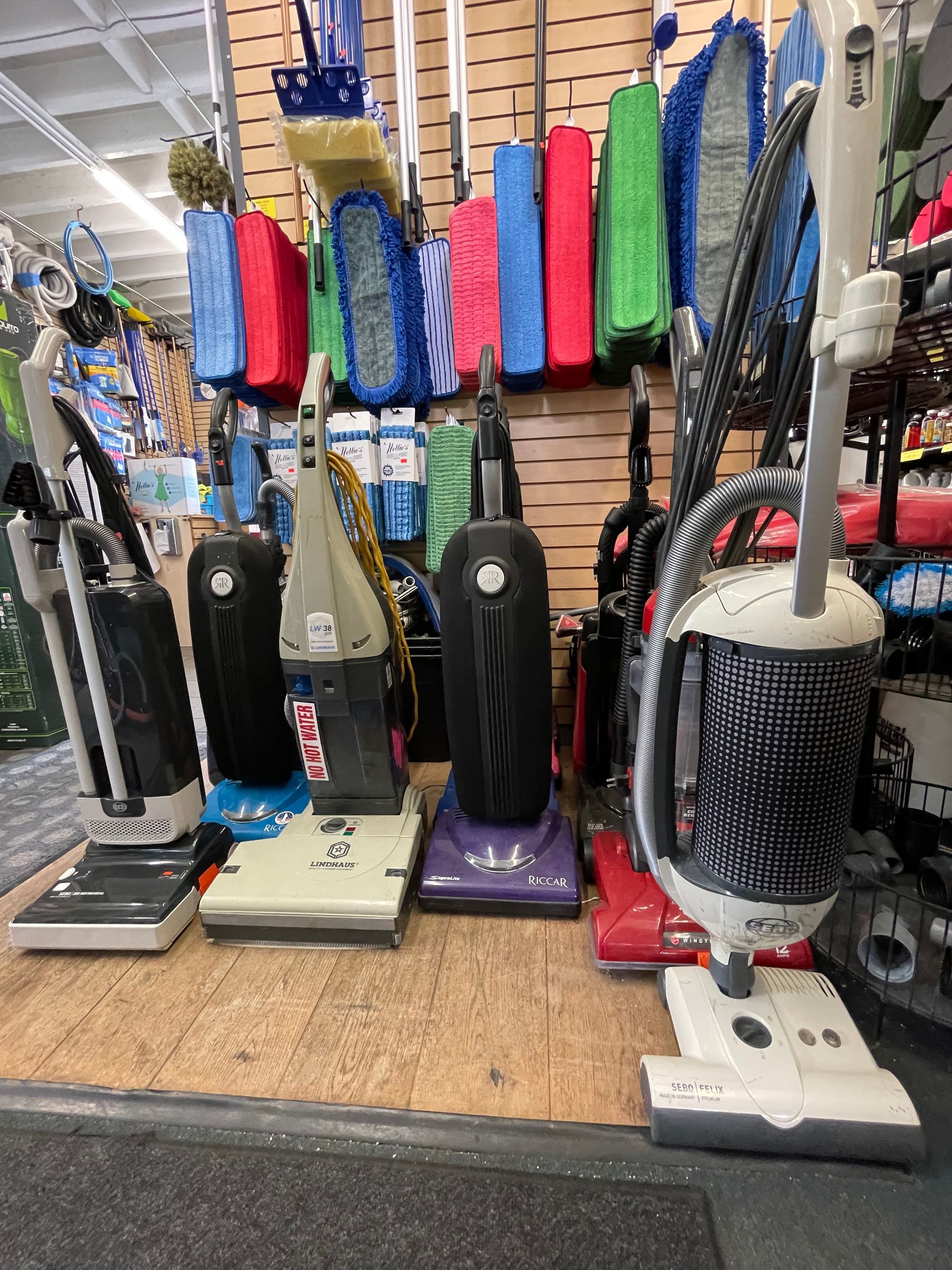 Row of upright vacuum cleaners on display in a store, with cleaning brushes hanging above.