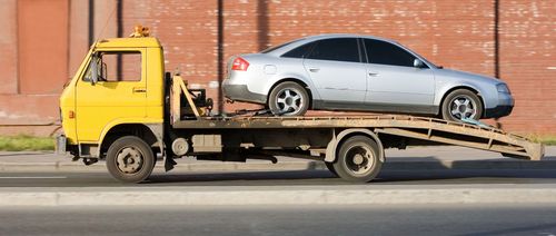 Yellow tow truck carrying a silver sedan on a flatbed trailer along a street.