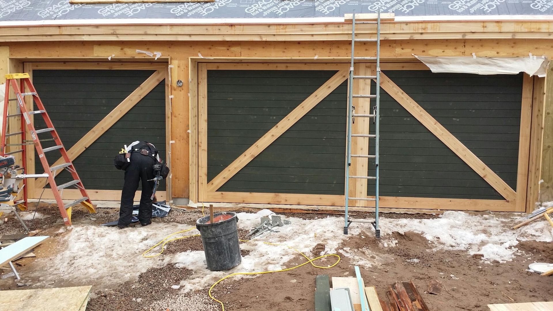 Exterior view of garage doors under construction with worker, ladder, and tools.