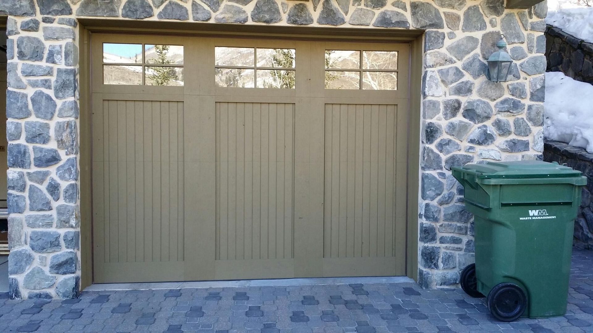 Tan garage door with windows and a stone exterior, green trash bin next to it.
