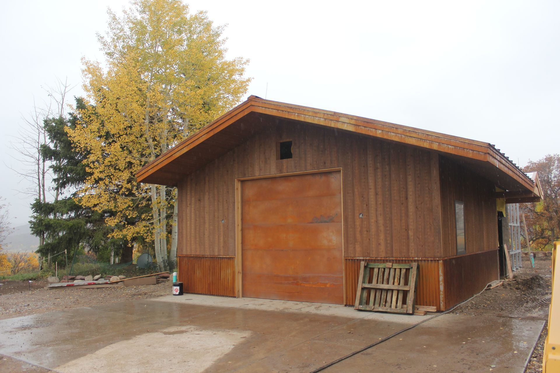 Wooden garage with brown door and roof; fall foliage in background.