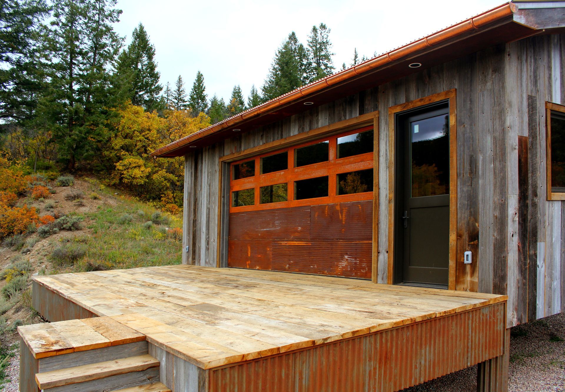Rustic cabin with a rusted garage door, deck, and steps against a hillside with trees.