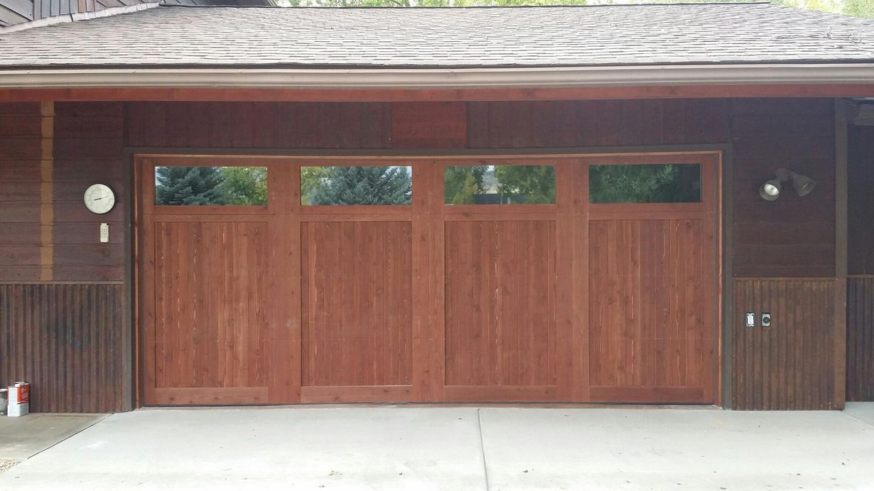 Brown wooden garage door with glass panes; set in a wood-sided building with a concrete driveway.