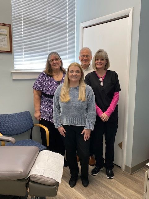 Four people stand in an office setting with a treatment chair in the foreground, posing for a group portrait.