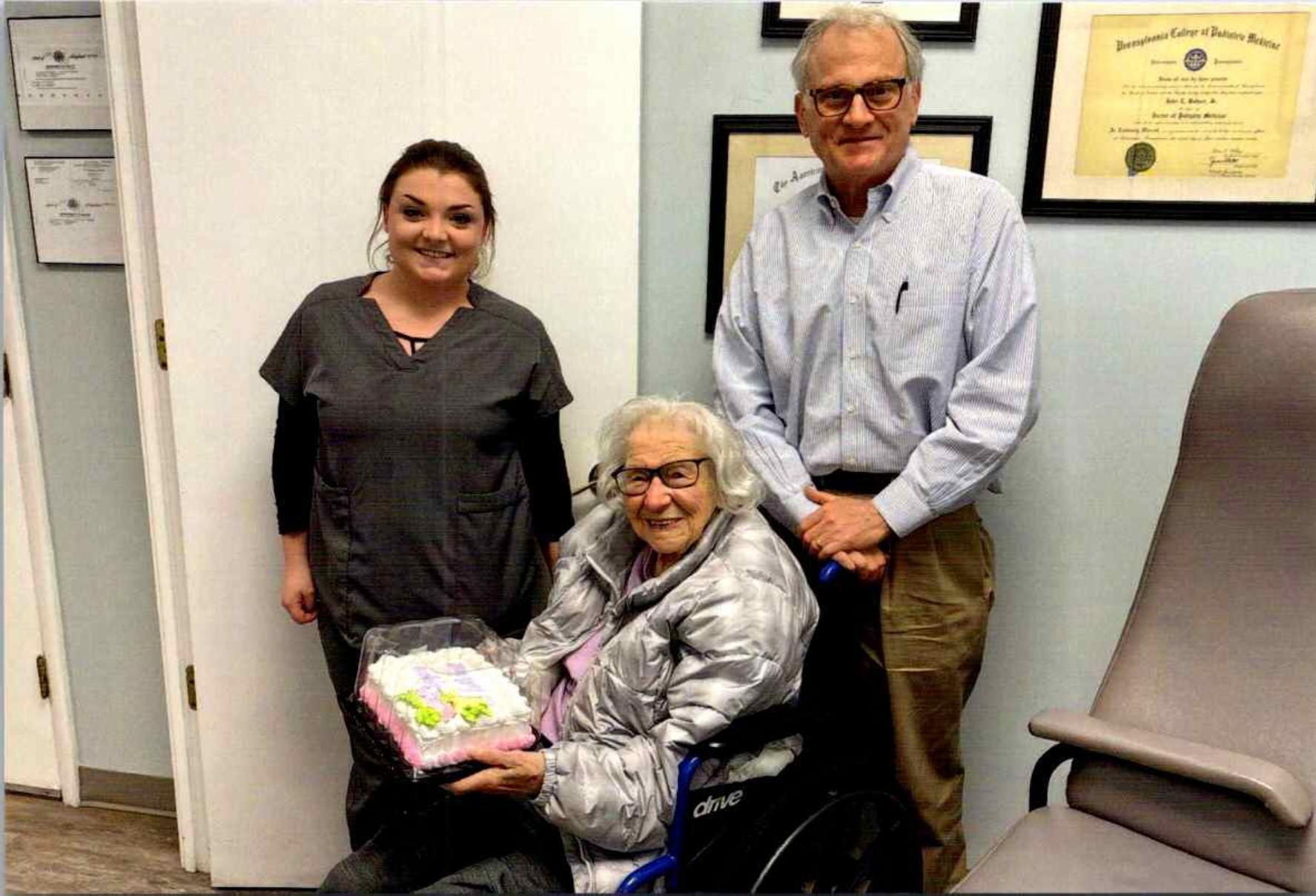 Three people are posing in an office, with a seated woman holding a gift basket.