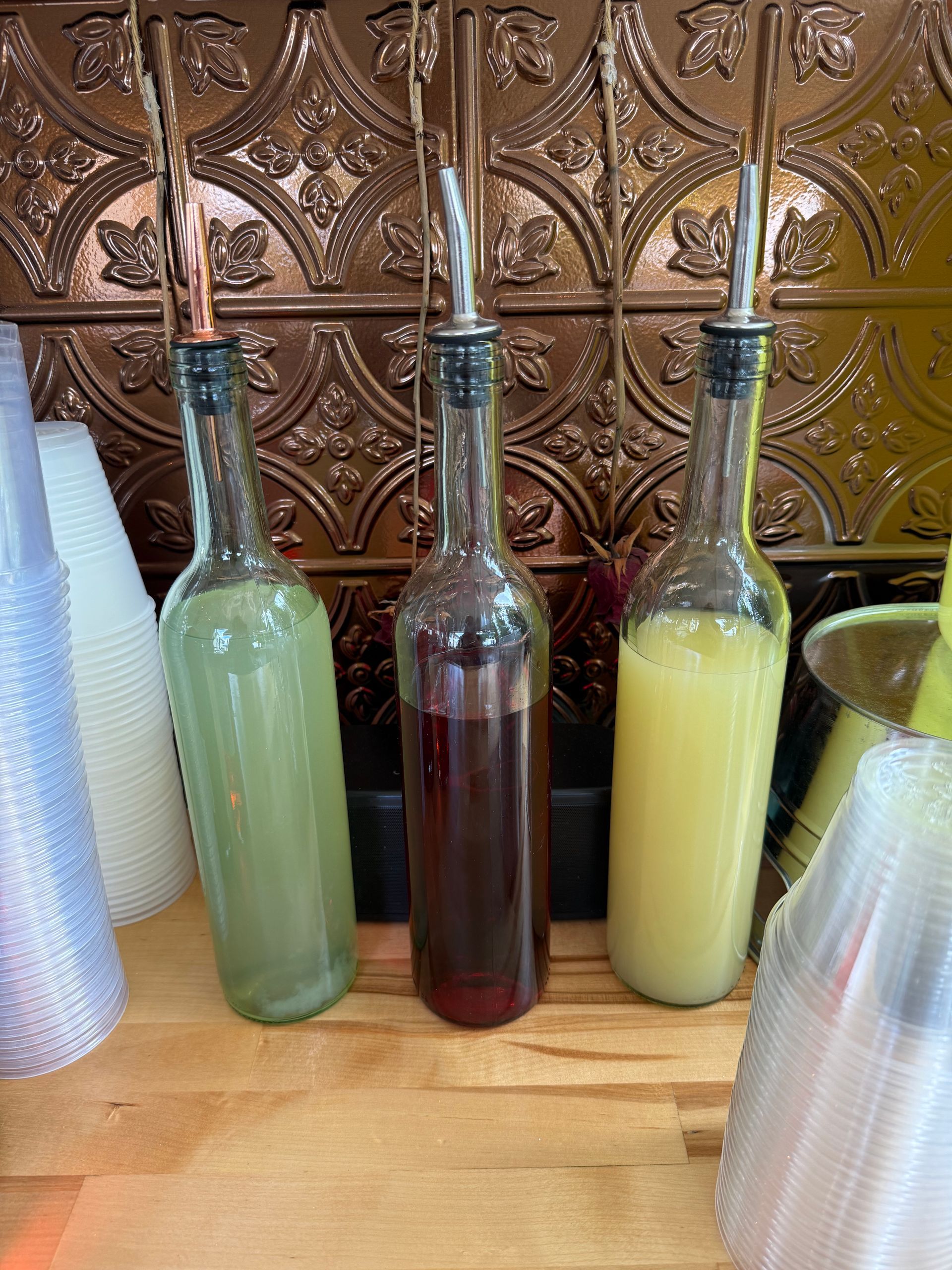 Three bottles of colorful syrups with pour spouts sit on a wooden surface against a textured copper-colored wall.