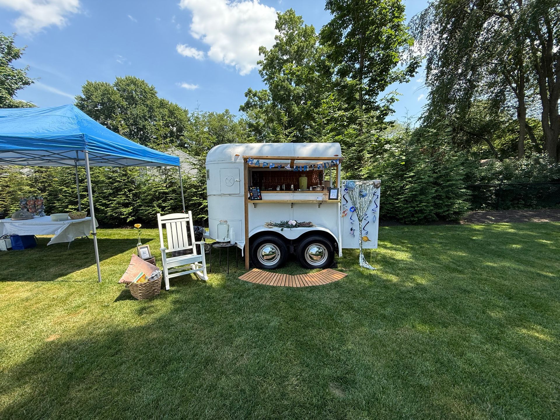 A white horse trailer converted into a bar sits on green grass under a blue sky. A blue tent and trees are in the background.