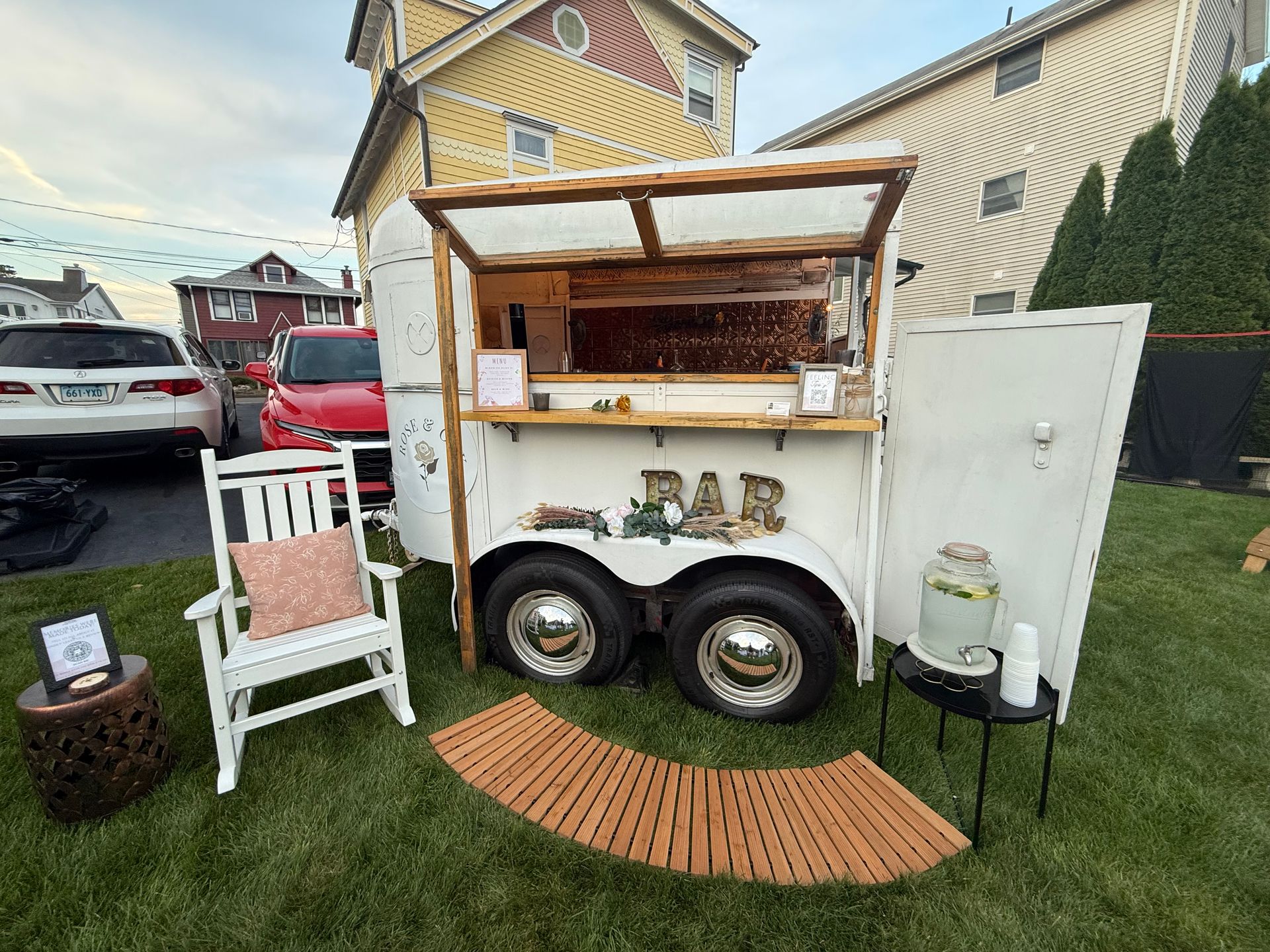 A white horse trailer converted into an outdoor bar with a rocking chair, small table, and refreshments. The bar is set up on a lawn.