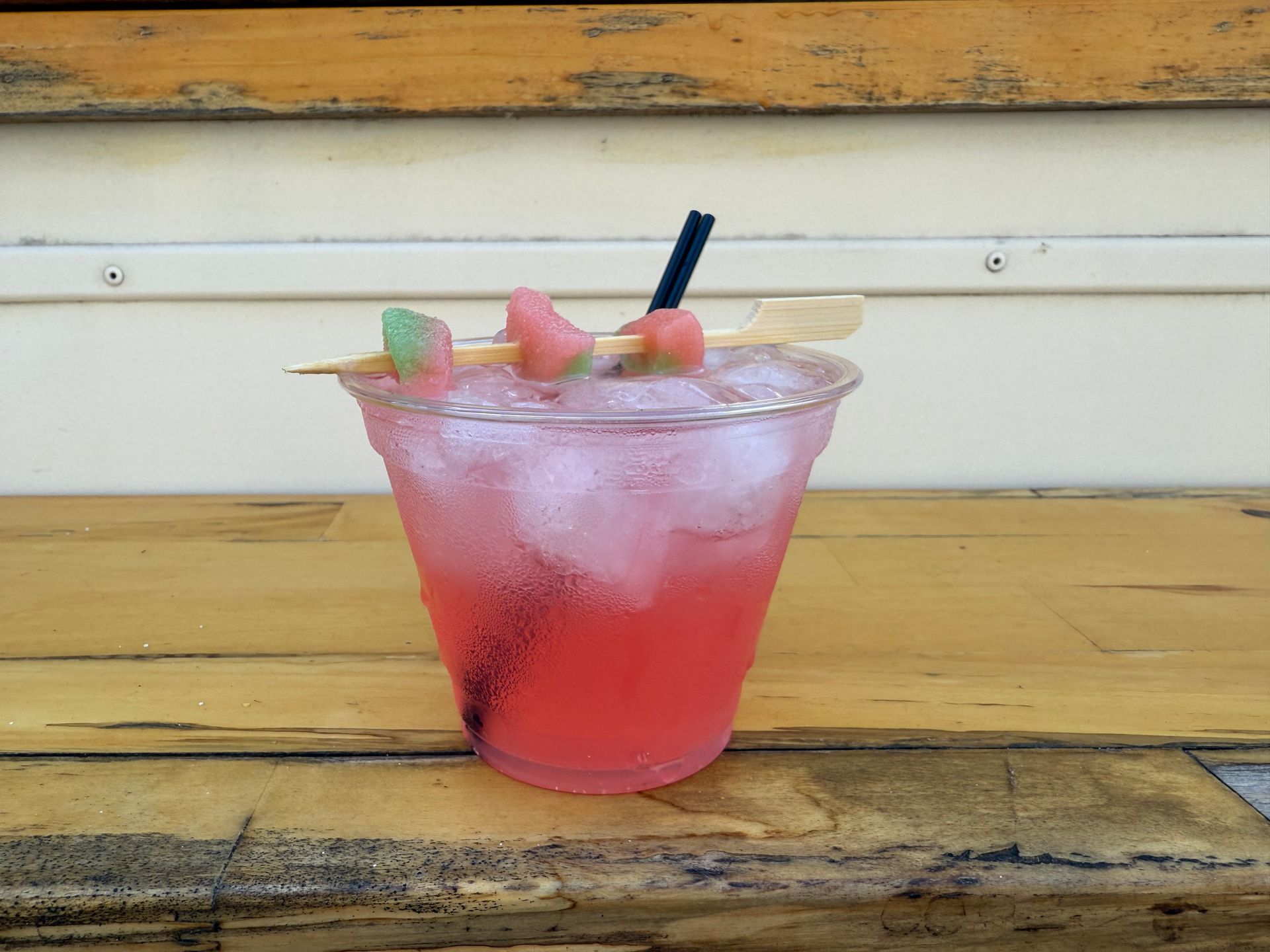 Watermelon cocktail in a clear plastic cup on a wooden surface, garnished with watermelon and a gummy candy.