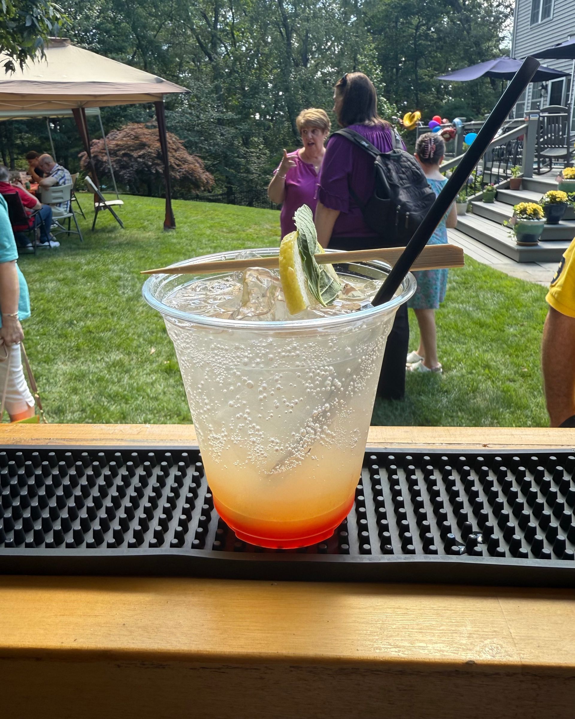 A cocktail in a plastic cup with a salt rim sits on a bar. People are in the background, enjoying an outdoor gathering.