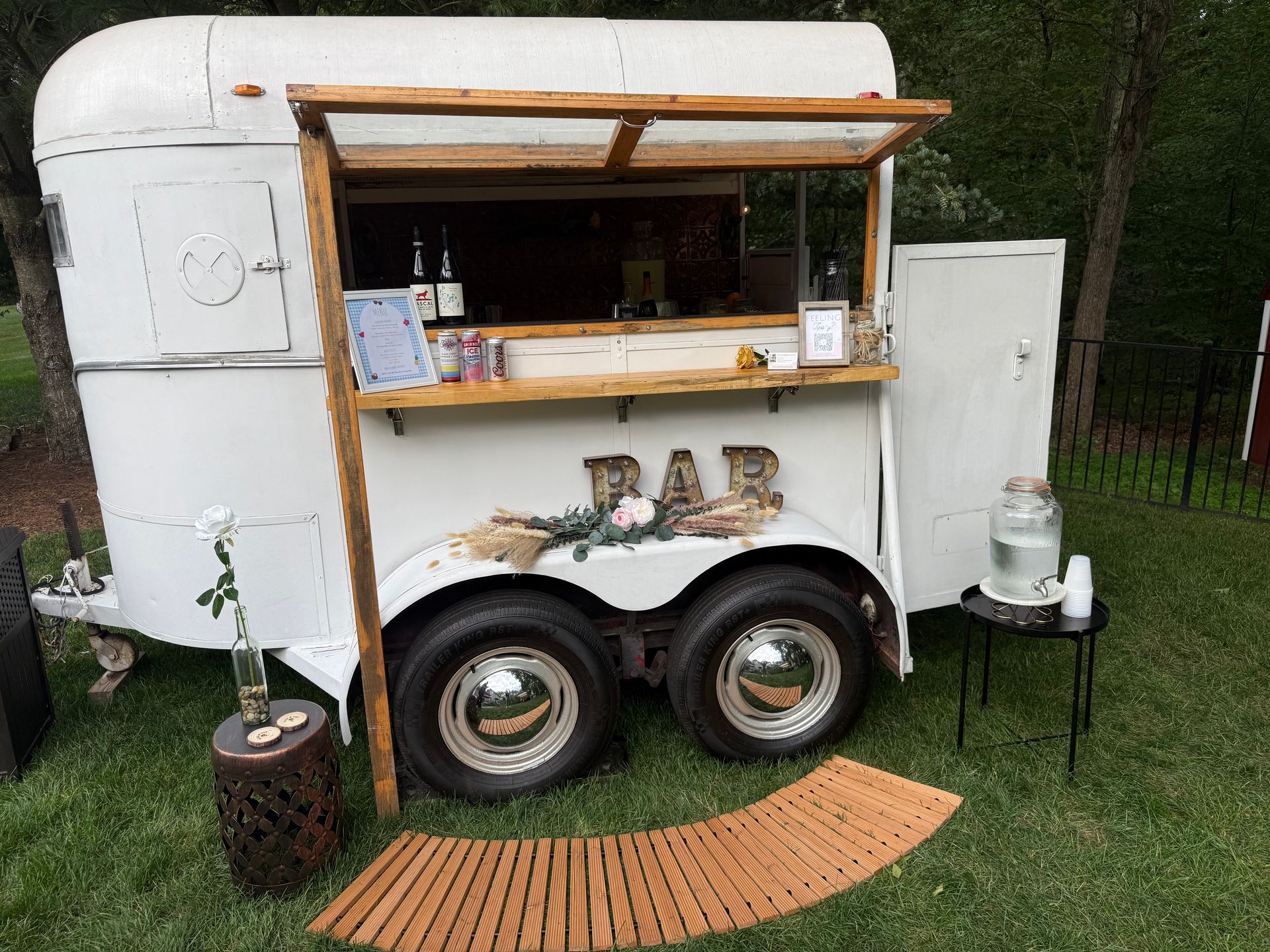 White horse trailer converted into an outdoor bar with wooden accents. The bar is set up on grass with a welcome mat and a water station.