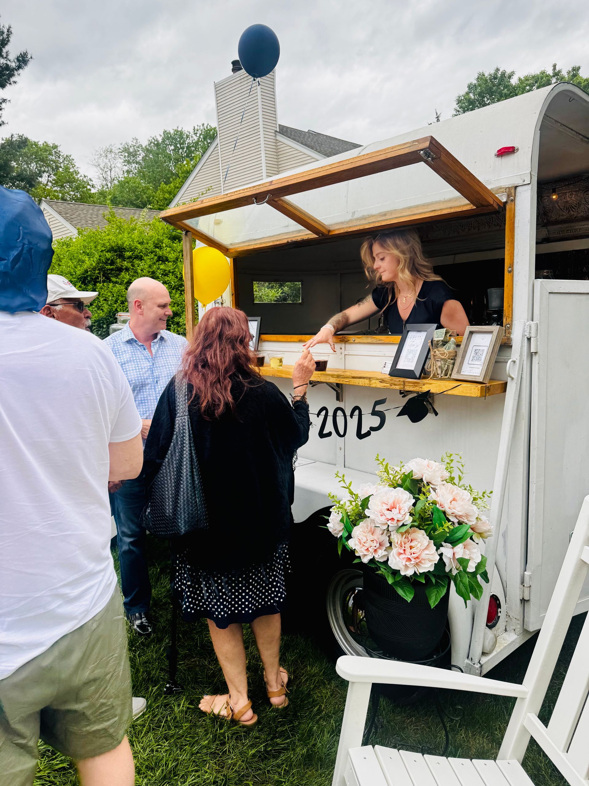 A woman serves from a mobile bar in a converted horse trailer. People queue outside on a grassy lawn; a flower pot sits nearby.
