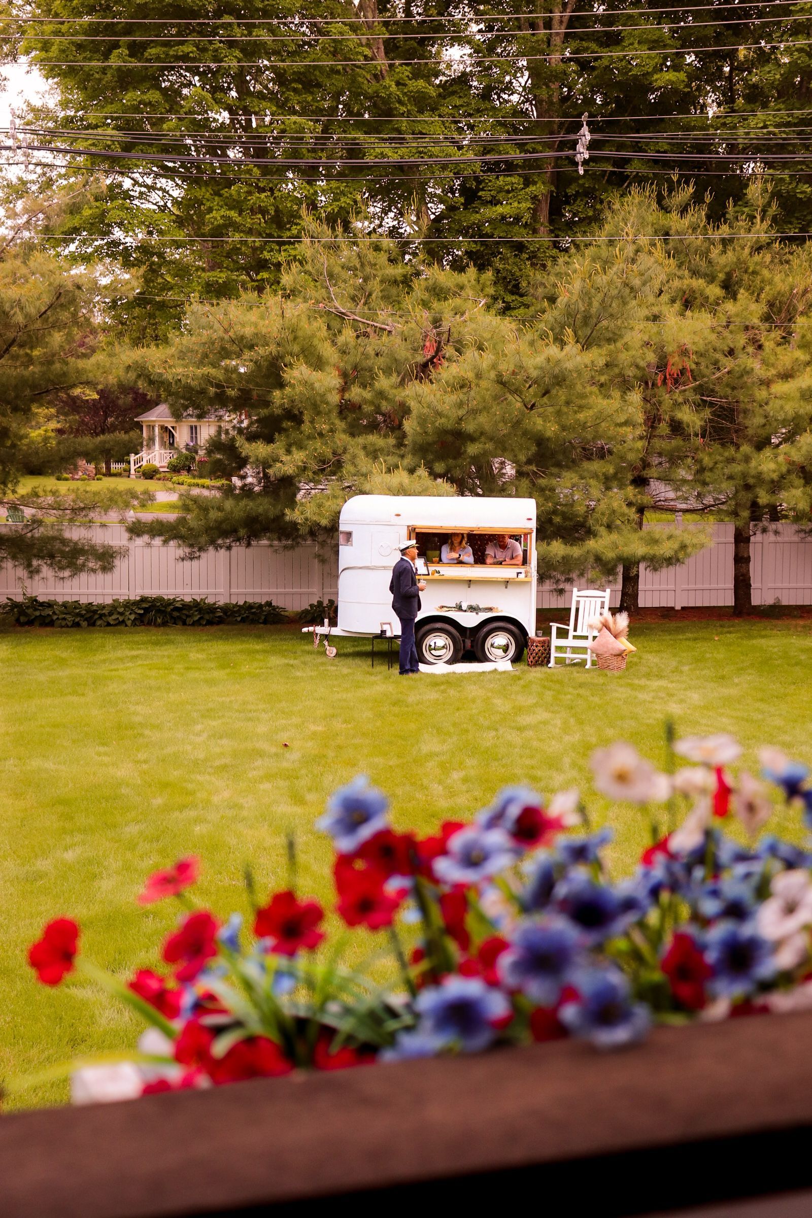 A food trailer in a grassy backyard with someone standing by it. Red, white, and blue flowers in the foreground.