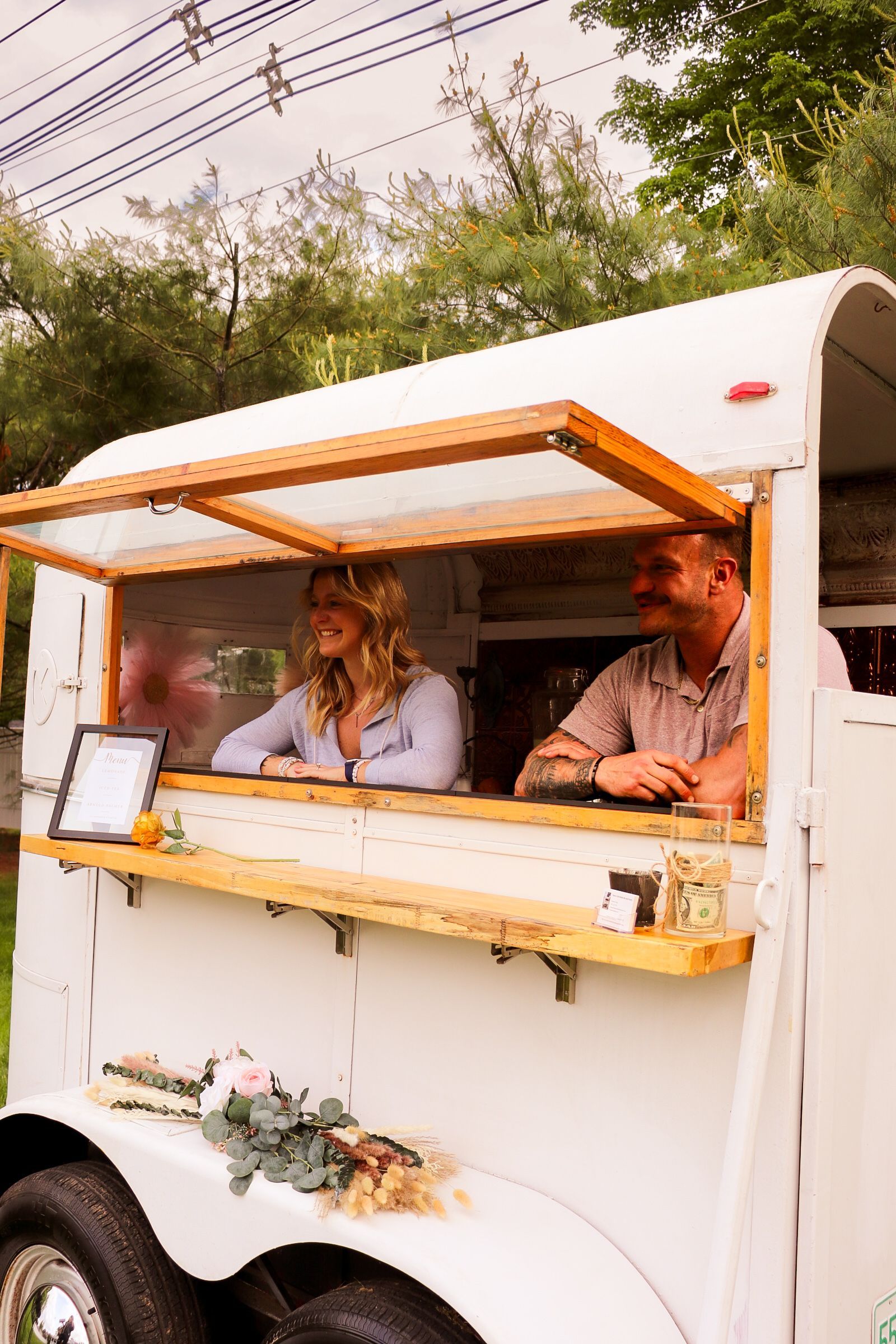 A white horse trailer converted into a bar, with a woman and man serving drinks.
