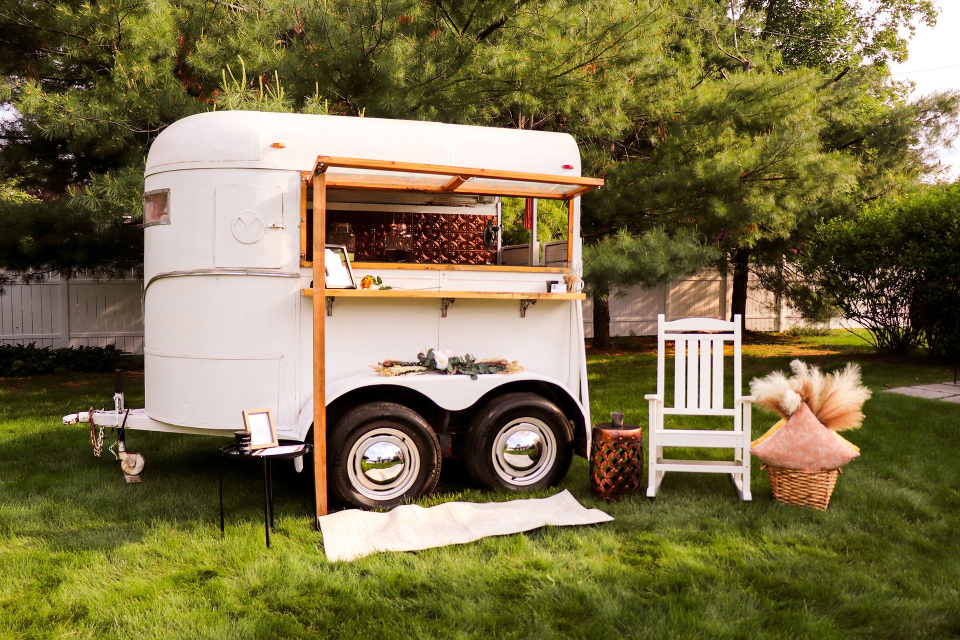 White horse trailer converted into a bar outdoors, with wooden awning, decor, and seating.