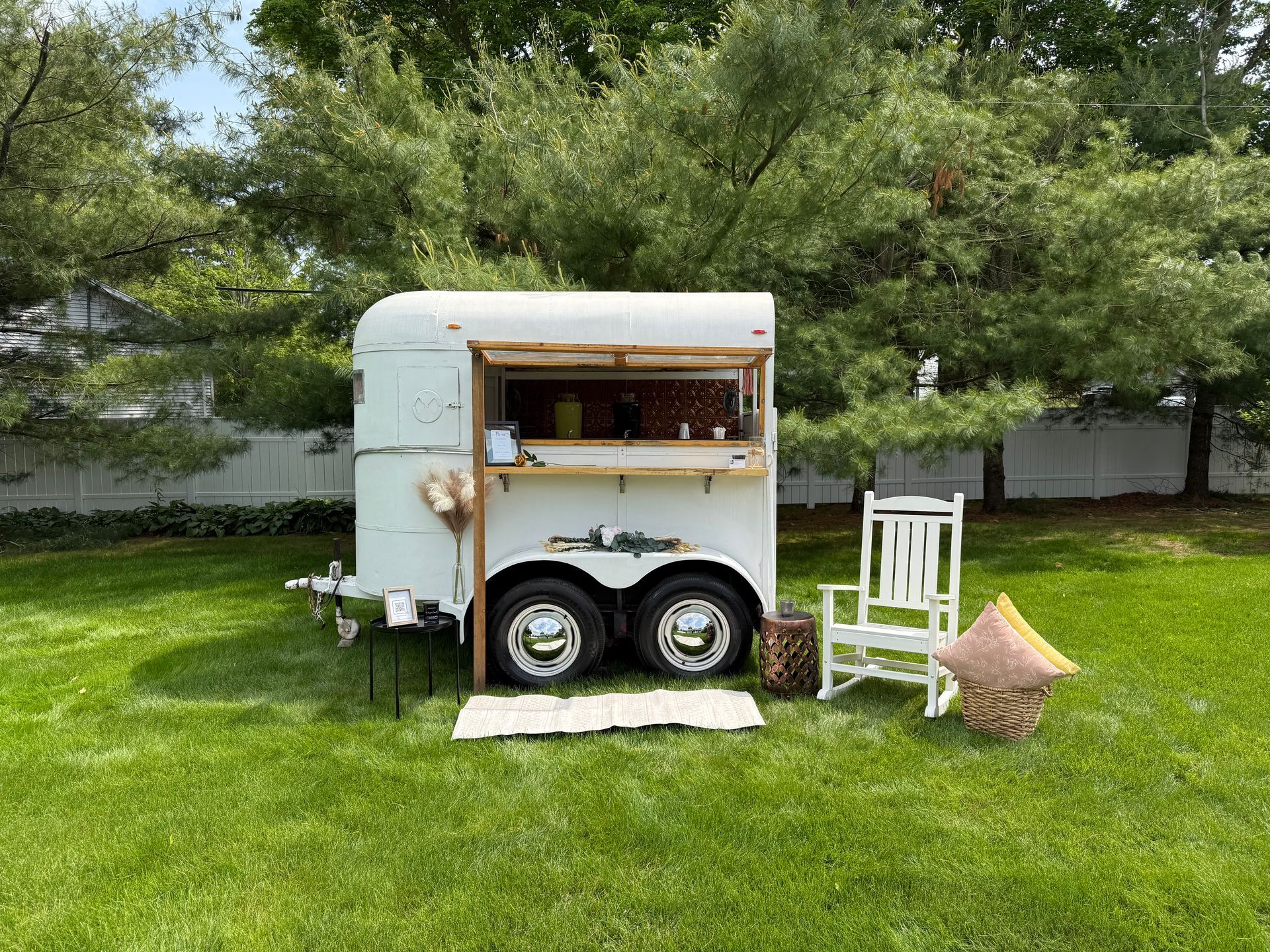 White horse trailer converted into a bar in a grassy backyard with a rocking chair.