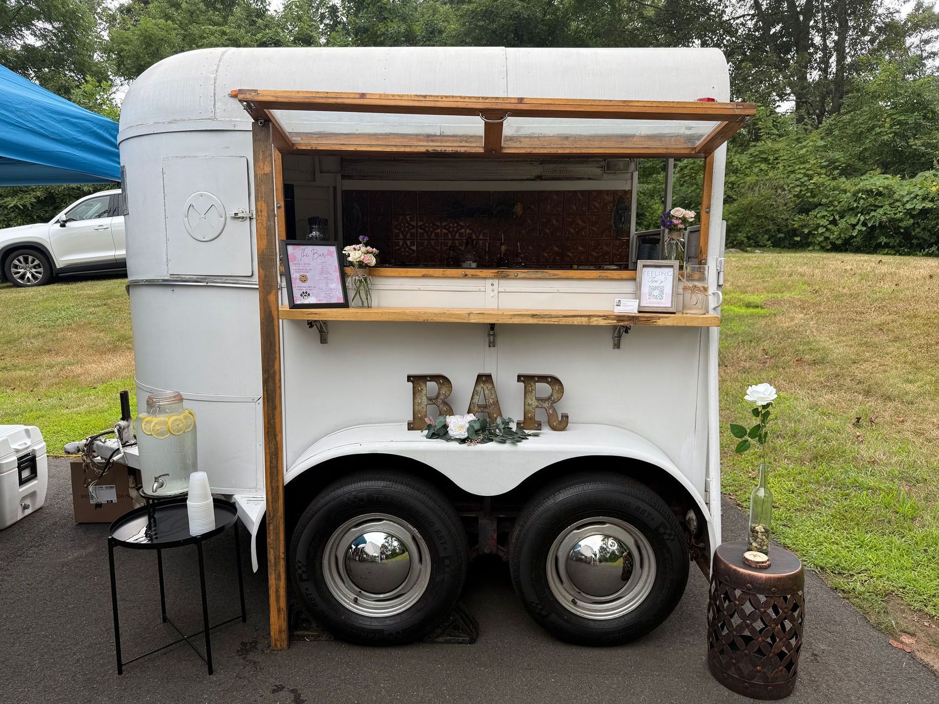 White horse trailer converted into a bar with wooden awning, wheels, and bar sign. A small table with lemonade and a decorative stool are beside it.