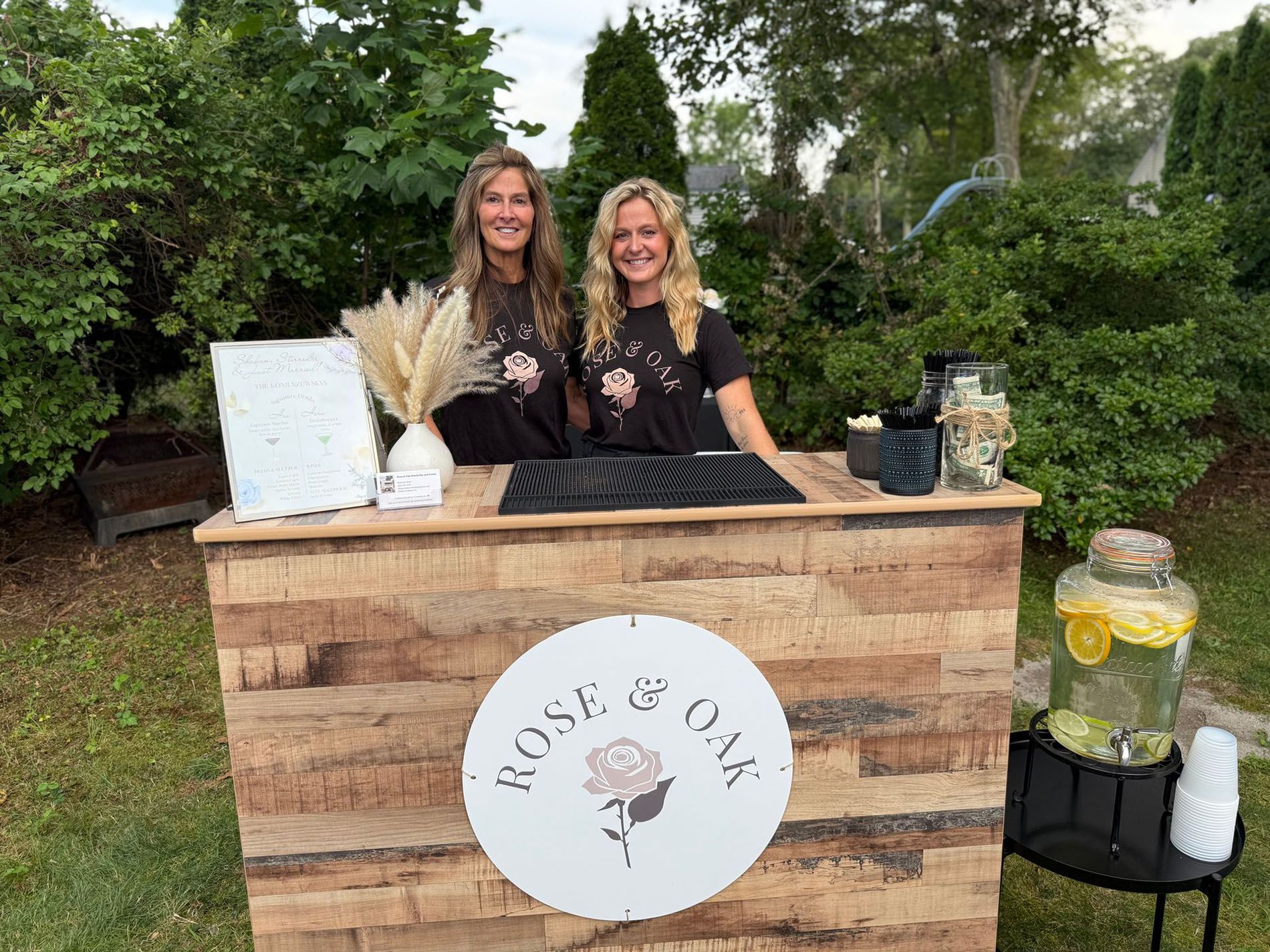 Two women at a wooden bar serving drinks outdoors. They smile, surrounded by greenery and a water dispenser.