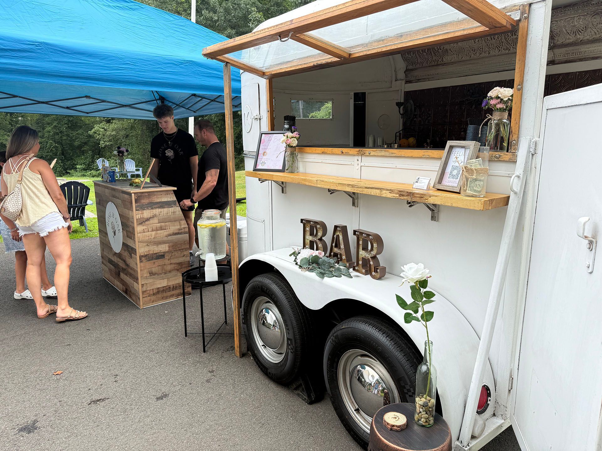 A mobile bar, a converted white trailer, with people ordering drinks outside.